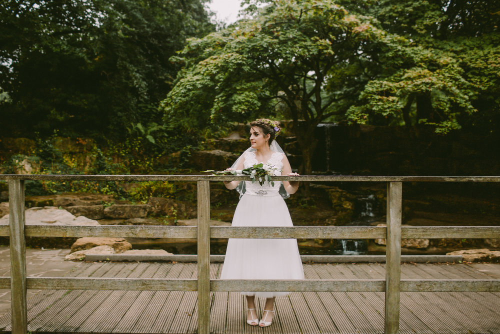 Bride with floral bouquet stood on bridge over pond.