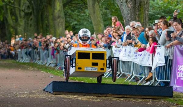 Soapbox vehicle racing over jump with crowds of people cheering.